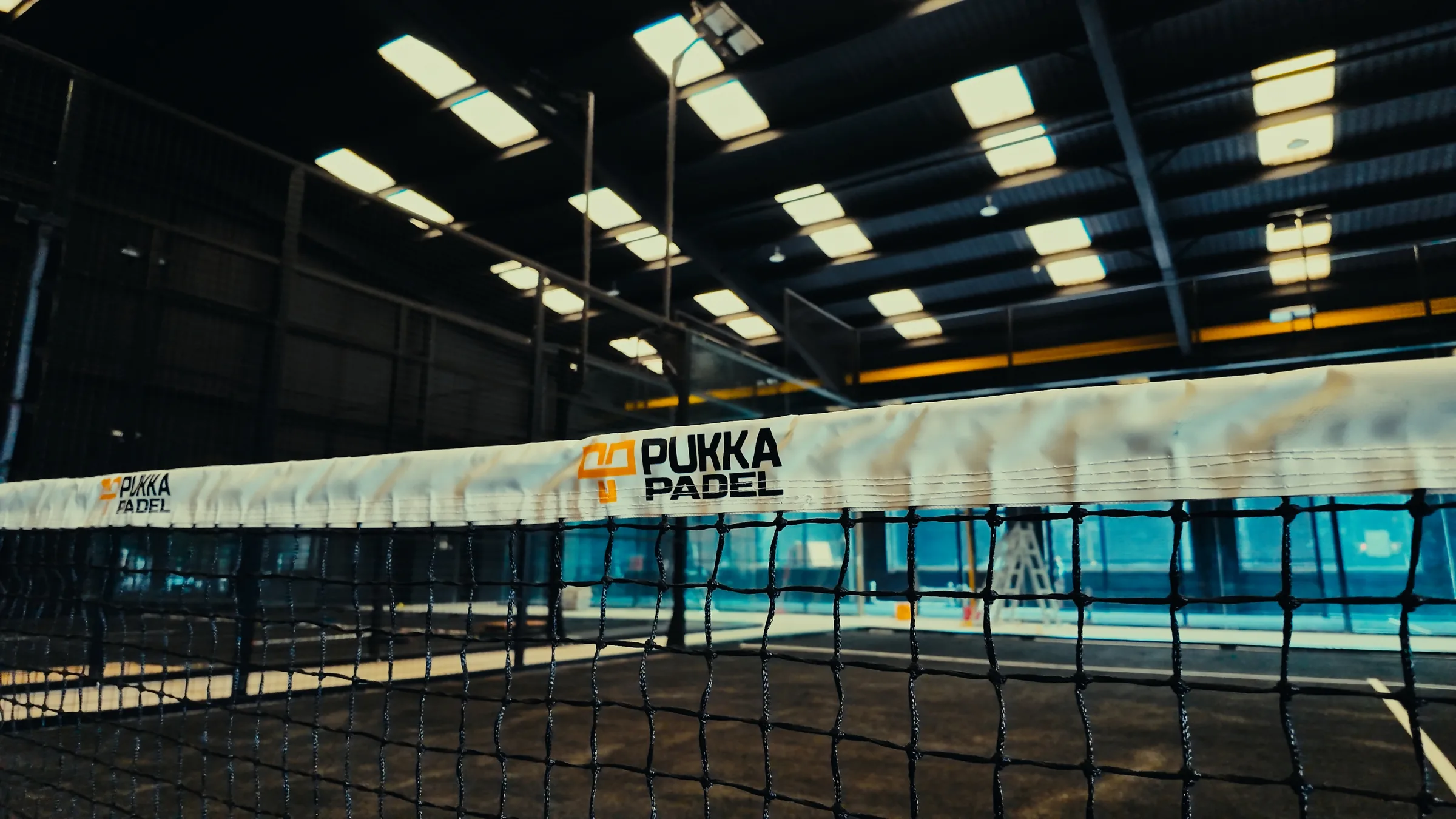 An indoor court at Pukka Padel Kidderminster with a branded net beneath a lit warehouse roof.
