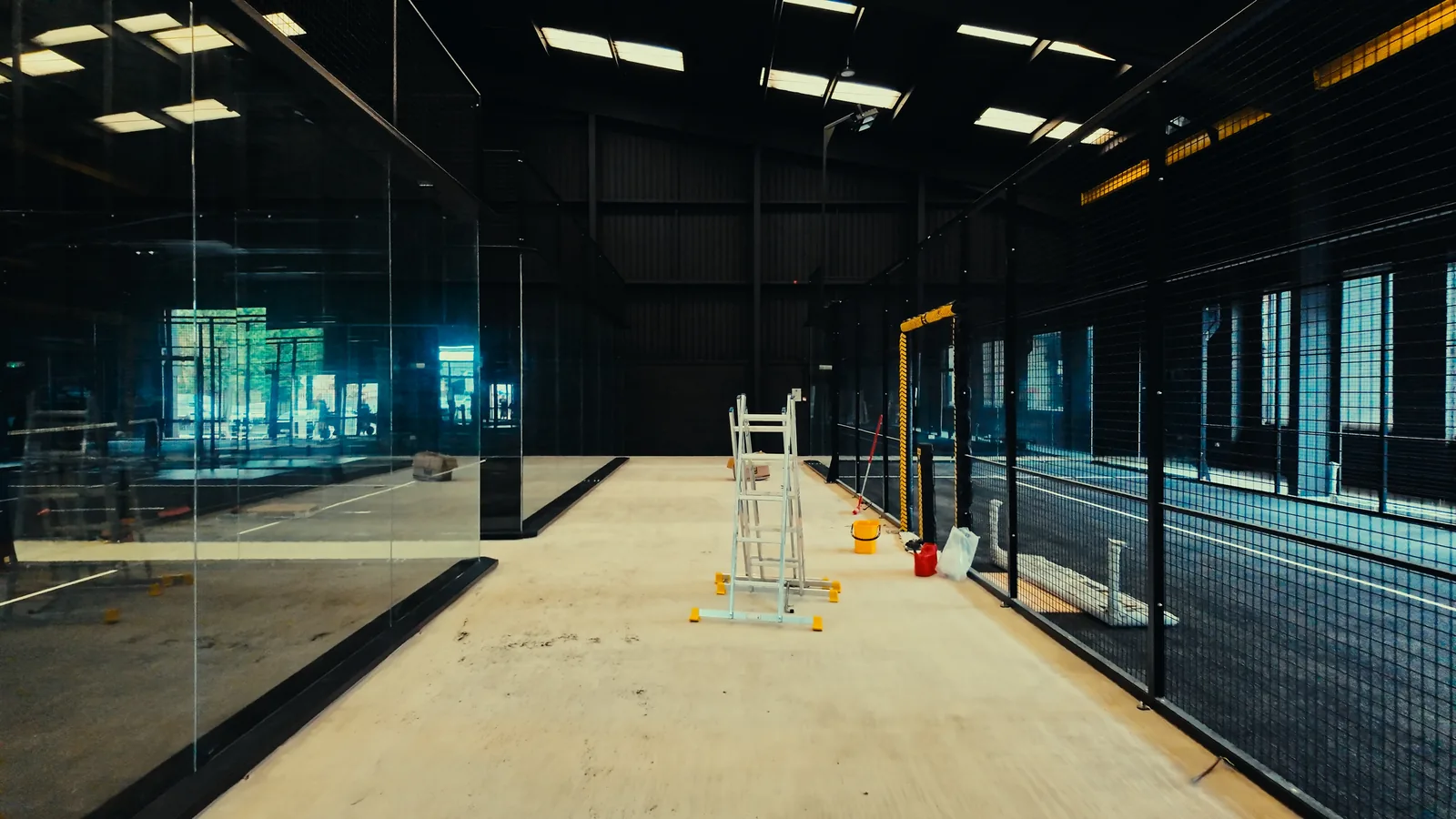 An indoor padel court going up: the glass back wall in its frame, the mesh along the side and the floor laid.