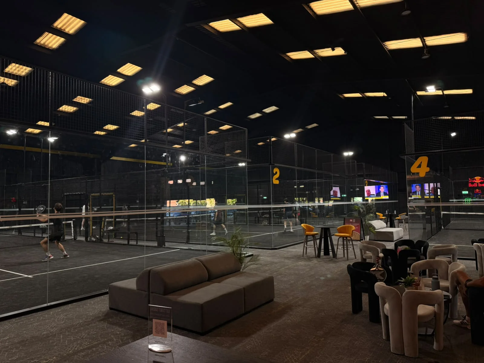 The player lounge at Pukka Padel Kidderminster at night, with the lit indoor courts beyond the glass.