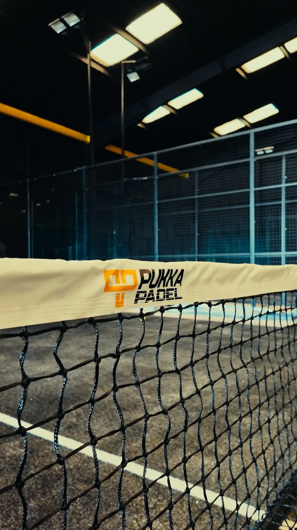 The net of a padel court close up, its band carrying the Pukka Padel mark, under the lights of an indoor hall.