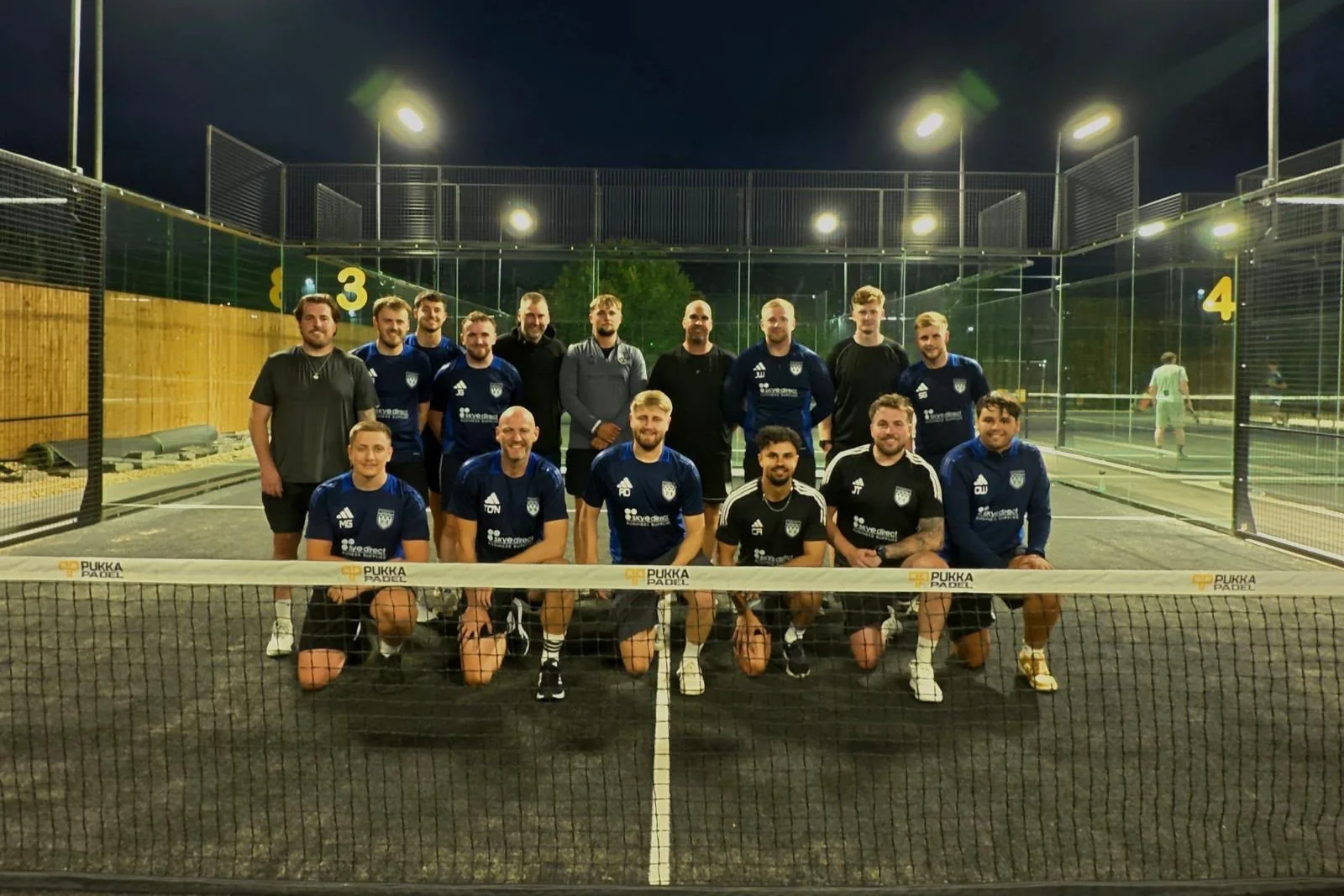 A company team lined up behind the branded net at Pukka Padel Leeds under the floodlights.
