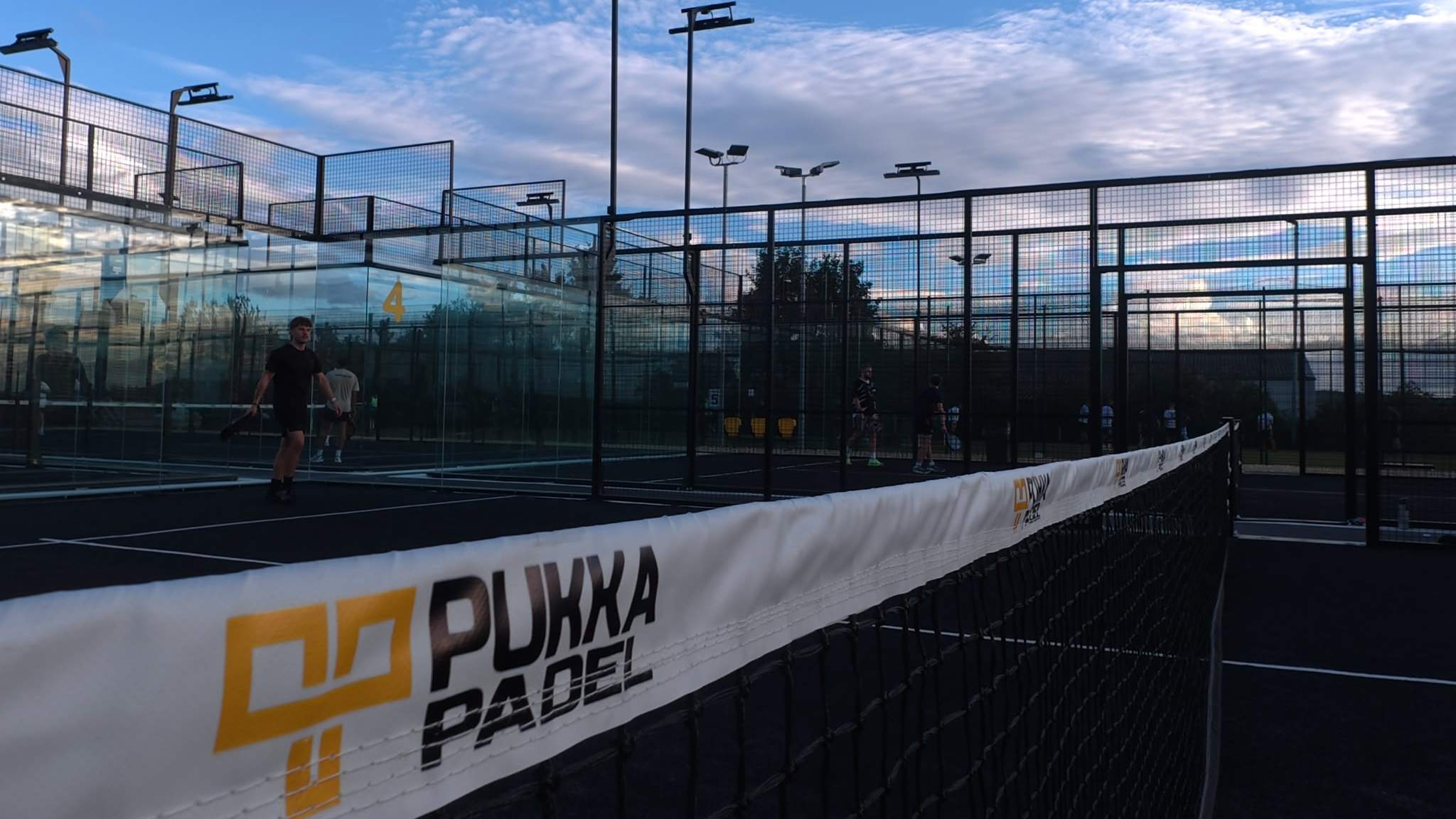 A Pukka Padel branded windbreak beside the outdoor courts at Pukka Padel Leeds under a blue sky.