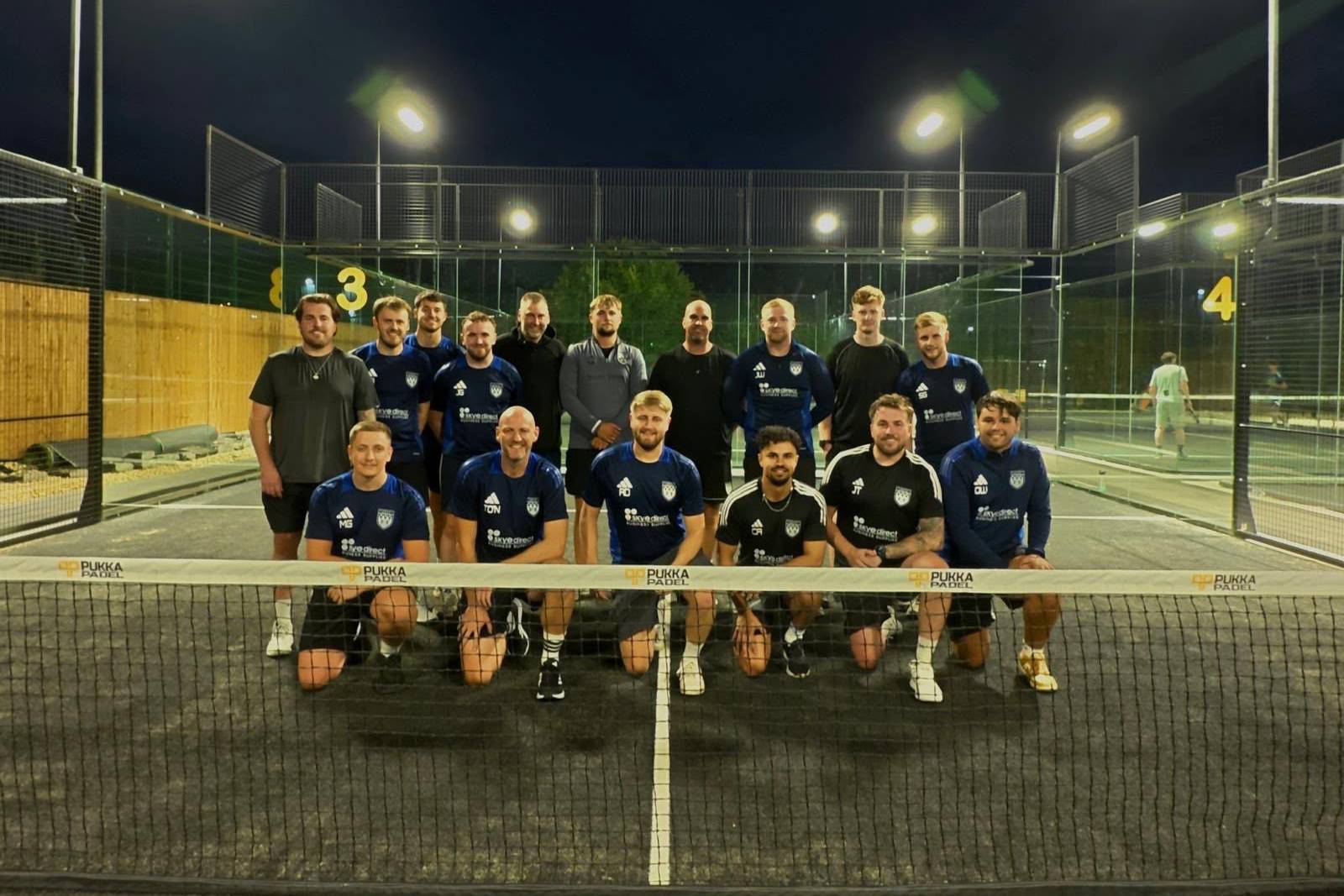 A company team on court at Pukka Padel Leeds at night, lined up behind the branded net under the floodlights.